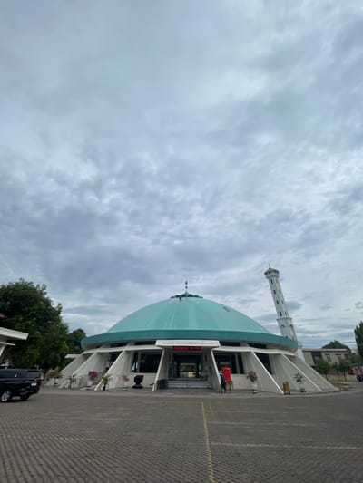 The image shows the exterior of a mosque with a large, teal-colored dome and a tall, white minaret. The mosque is situated in a paved courtyard. A black SUV is parked to the left of the mosque. A person in a red shirt is visible near the entrance of the mosque. The sky is overcast with gray clouds. The text on the mosque reads "MASJID RAUDHATURRAHMAN" and "Senyuman Bersama".