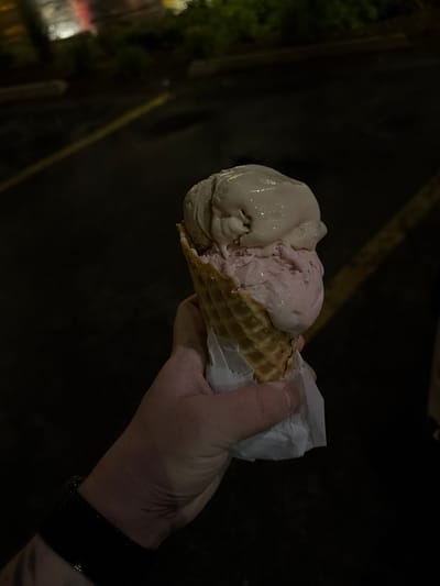 A person's hand, wearing a black watch, holds a waffle cone with two scoops of ice cream, one pink and one light brown, on a dark background. The background appears to be an outdoor setting at night, possibly a parking lot, with faint yellow lines visible on the ground and some blurred greenery in the distance. The lighting suggests artificial light sources are present. The image was taken in Arlington Heights, USA.