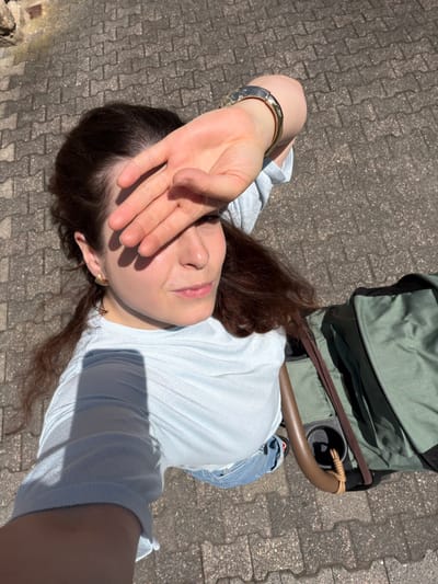 The image displays a young adult female outdoors under direct sunlight. She has long, wavy, dark brown hair and fair skin with visible freckles. She is wearing a light blue ribbed long-sleeved top and blue denim bottoms, partially visible. Her left wrist is adorned with a hammered gold-colored cuff bracelet, and her right ear features a gold-toned, irregularly shaped stud earring. Her left hand is raised, shielding her face from the sun and casting a prominent shadow across her eyes and upper face. Her head is tilted slightly to the right, and her gaze is directed upwards and to the right. The ground is paved with interlocking gray concrete pavers. In the upper left background, a portion of a brick structure is visible. To the right of the woman, a green fabric object, consistent with a stroller component or large bag, is present. It features a brown handle, an integrated black cup holder, and a light brown braided cord wrapped around the handle. No specific visual markers are present within the image to determine a precise location within Schmittweiler, Germany.