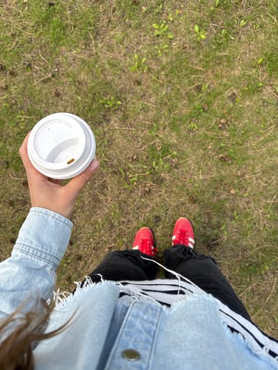 The image shows a person standing on grass, holding a white disposable coffee cup in their right hand. The person is wearing black jeans with white stripes on the side, a light blue denim jacket and red sneakers with white stripes. The grass appears to be a natural setting. The location is not identifiable as Chelsea, Canada from the image.