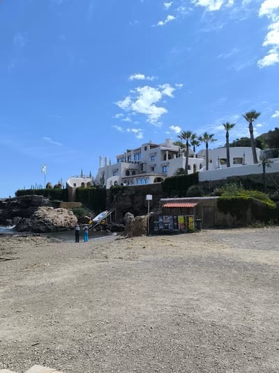 The image depicts a coastal scene in Sant Joan de Labritja, Spain, under a partly cloudy sky. A large, white building complex with multiple levels and rounded architectural features stands on a rocky cliff overlooking a beach. Palm trees are scattered around the complex. Below the building, on the beach, there are two people standing near a small boat resting on a ramp. A small structure with a red awning and posters on display is situated near the beach. The foreground is dominated by a gravelly, sandy area. The ocean can be seen lapping against the rocks along the shoreline.