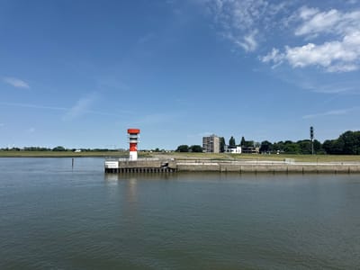 The image captures a wide view of the Elbe River in Brunsbüttel, Germany, on a clear, sunny day with a few scattered clouds in the bright blue sky. The foreground is dominated by the dark, rippling water of the river.

On the left side of the image, a red and white lighthouse stands on a concrete pier. The lighthouse has a circular base and tapers upwards, topped with a red section. A white sign with black text is affixed to the pier near the lighthouse. The pier itself is constructed of concrete with wooden pilings extending into the water.

Beyond the pier and lighthouse, the bank of the river is visible, featuring a grassy area with a paved walkway. In the middle ground, several buildings are present, including a tall, multi-story residential building with a gray facade. Further to the right, a cluster of trees and a communication tower are visible. In the distance, wind turbines can be seen on the horizon, indicative of the industrial and coastal nature of the region. The overall scene is one of a working port or waterway with both navigational aids and urban development.