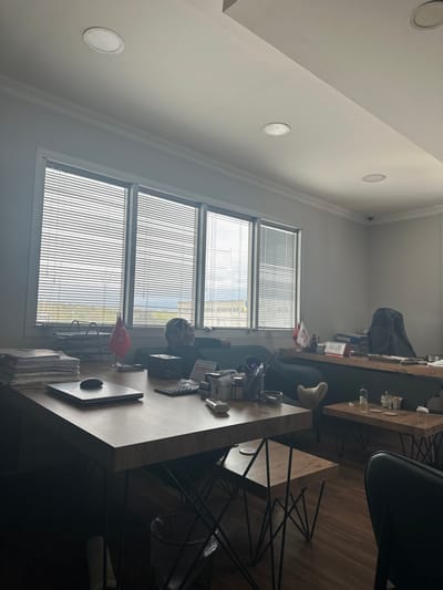 The image depicts an interior office scene located in Kemalpaşa, Turkey. A male individual is observed reclining on a dark-colored sofa positioned against a wall featuring multiple large windows. The windows are fitted with horizontal blinds, partially obscuring a view of distant buildings and an overcast sky. The man wears a dark top and has a clear reflective visor or face shield pushed up onto his head; his eyes are directed upwards.

In the foreground, a rectangular wooden desk occupies a significant portion of the frame. On its surface, from left to right, are a tall stack of papers, a closed silver laptop with a black computer mouse positioned next to it, a miniature Turkish flag, a calculator, a pen holder containing multiple writing instruments, various small containers, an ashtray, and a lighter. Below this desk, a small black waste bin is visible.

To the right and further back, another wooden desk is present, cluttered with binders and documents. A miniature Peruvian flag is prominently displayed on this desk. A dark office chair is partially visible behind it. Two smaller, square wooden tables with geometric black metal bases are situated between the foreground desk and the back desk. One of these tables holds a clear bottle, two small glass bowls, and round coasters.

The room features white walls and a white ceiling with multiple recessed circular light fixtures. The flooring is made of dark wood. The overall lighting suggests ambient natural light entering through the windows, creating some glare and shadowed areas within the room. No active interactions are depicted.