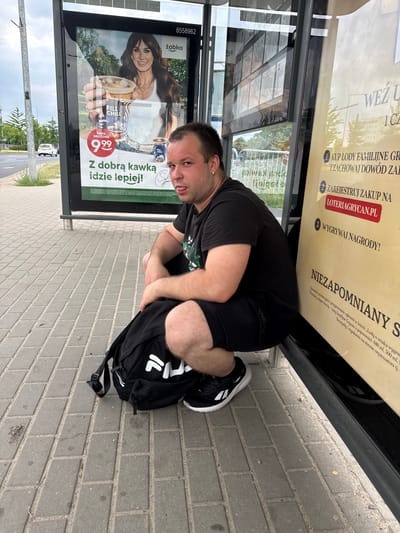 A young adult male is squatting on a paved sidewalk next to a bus stop shelter in Poznań, Poland. He is wearing a black t-shirt and black shorts, and a black backpack with the FILA logo is on the ground beside him. The man is sticking his tongue out towards the camera. Behind him, a large advertisement panel is visible, featuring a woman holding a cup of coffee and promoting a product from the "Żabka" convenience store chain. Another advertisement panel to the right, with text in Polish, advertises a lottery promotion for "Grycan" ice cream. The environment appears to be an urban street with a road and sidewalk, indicating a public transportation stop.