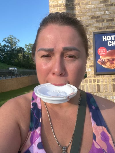 A medium shot captures a woman from the chest up, holding a white paper bowl with whipped cream in her mouth. She is outside, with a brick wall and a sign for "HOT CH..." in the background, suggesting a fast-food establishment. To the left, a green lawn leads to a fence and a road with a vehicle, indicating a suburban or highway-adjacent location. The sky is a clear blue. The woman has brown hair, dark eyebrows, and is wearing a colorful patterned top. A silver chain necklace is visible. The image was taken in Emerson, United States of America.
