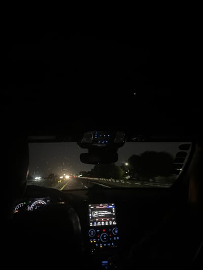 A nighttime scene is observed from inside a moving vehicle on a road in Celaya, Mexico. The perspective is from behind the driver, whose left shoulder and arm are visible gripping the steering wheel. The dashboard gauges are dimly illuminated. A central infotainment screen displays music information, specifically "Flor de Maria" and "La Chica Del Bikini" by Luis Miguel. Below the screen, illuminated controls are visible. An overhead console with blue illuminated buttons is mounted above the rearview mirror. The windshield exhibits faint streaks, suggesting moisture. Outside the vehicle, a multi-lane road is visible with numerous distant white and yellow headlights from oncoming traffic and red taillights from vehicles traveling in the same direction. Dark outlines of trees and foliage line the right side of the road under a dark, overcast sky. A subtle silhouette on the far right of the frame suggests the presence of a front passenger.