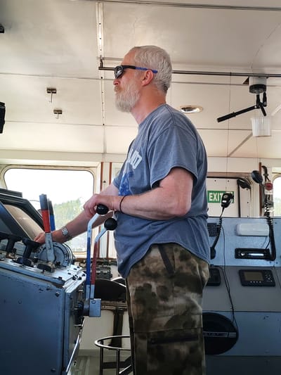 A man with gray hair and a beard, wearing sunglasses with blue frames, a gray T-shirt, and camouflage pants, stands in profile inside a ship's control room, likely in Chaika, Russia. He is actively piloting the vessel, with both hands engaged on a grey control console. His left hand is on a smaller lever, while his right hand grips two prominent black-knobbed levers, suggesting he is managing speed or direction.

The functional control console is equipped with multiple levers, buttons, and communication equipment, including microphones with coiled cords. A small digital display showing "VEGA" and a radio unit are also integrated into the panel. Bright daylight enters through large windows on the left and front, indicating a clear, sunny day. A green sign with "EXIT" is visible in the background on the right. The overhead ceiling is white and paneled, featuring hooks and a small white bucket suspended by a pole. The scene captures the focused activity of navigating a maritime vessel.