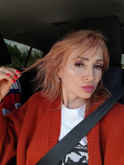 The media file shows a woman with pinkish-blonde hair and makeup, sitting in the passenger seat of a car. She is wearing an orange knitted cardigan over a white t-shirt with a black lace-like pattern. A black seatbelt is visible across her chest. Her left hand is raised, and she is holding a strand of her hair. Her nails are painted red. The background suggests an outdoor setting with trees visible through the car window. The lighting indicates it is daytime. The woman's expression is a neutral gaze towards the camera, with slightly parted lips. No visible text is present in the image. The overall scene depicts a woman in a car, possibly on a journey.