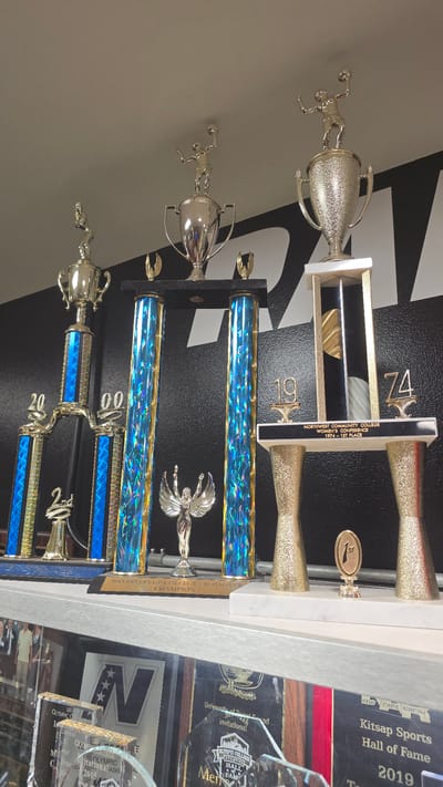 This image shows a collection of trophies on a shelf in what appears to be a sports facility or a school in Bremerton, United States of America. The trophies vary in size and design, with several tall, multi-tiered trophies featuring blue and gold cylindrical columns. Some trophies have figures of athletes on top, including one that appears to be a basketball player. There are also smaller trophies and plaques visible. In the background, a black wall with white graphic designs is visible. Below the shelf with the trophies, other items, possibly more awards or photographs, are visible on another shelf. The lighting is overhead, creating some glare on the trophies.