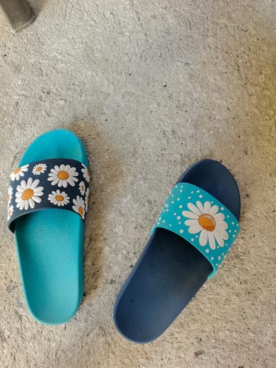Two flip-flops are lying on a textured concrete surface. The flip-flop on the left is turquoise and has a navy blue strap adorned with white daisies with orange centers and smaller white dots. The flip-flop on the right is navy blue and has a turquoise strap decorated with a large white daisy with an orange center and small white dots. The scene appears to be outdoors, possibly in a yard or on a sidewalk in Novoselivka, Ukraine, given the concrete ground and lack of indoor elements. There are no people or animals present in the image. The lighting suggests it is daytime, with shadows cast on the ground.