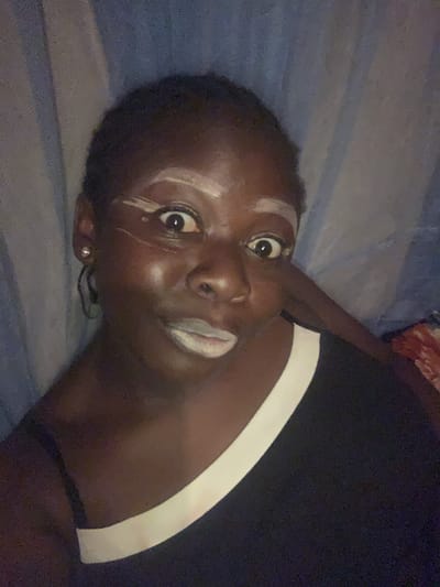 An image captures a person, appearing to be lying down or reclining, their head tilted significantly to the left. The subject is an individual with dark skin and short, dark hair, positioned centrally in the frame. Their gaze is directed intently at the camera, with wide-open eyes that convey a sense of surprise or intense focus.
A distinctive feature is the white or light-colored substance applied to their face. There are prominent white marks above and below their right eye (viewer's left), extending across the brow and cheekbone. Additionally, their lips are covered with the same white substance. In their earlobe, a small, light-colored stud earring is visible.
The person is wearing a dark-colored top, possibly a t-shirt, which features a wide white stripe running diagonally across the shoulder and upper chest area on the right side of the frame. The background consists of a soft, textured fabric in muted blue and grey tones, likely bedding or a blanket, suggesting an indoor setting. The lighting is dim, casting soft shadows.
The scene appears to be a direct portrait or selfie, potentially for artistic expression, a playful act, or a cultural demonstration given the facial markings. There is no visible text or specific time of day indicated beyond the low light conditions. The location context provided is Kukum, Nigeria.