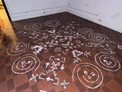 A room with a checkered brown floor displaying artwork. White chalk drawings depict spirals with geometric shapes inside, along with isolated triangles, squares, and cruciform figures. Small pebbles are arranged in clusters and lines around and sometimes within these chalk symbols. The walls are white with electrical outlets, adding a modern element to this floor-based art installation.