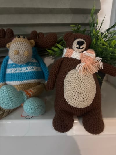 The image depicts an indoor scene in the city of Kozlovo, Russia, featuring two crocheted plush animal figures arranged on a white, flat surface. To the left, a light tan crocheted moose or reindeer-like figure wears a blue and white striped sweater. Its dark brown antlers are partially visible behind its head, and two detached light blue spherical elements, possibly feet, rest near its base. To the right, a dark brown crocheted bear-like figure stands upright, adorned with a light tan oval-shaped belly patch and a striped scarf in white and peach tones around its neck, tied with tassels. Behind the figures, a potted artificial plant with green leaves is visible, along with a light gray, horizontally ridged background element, possibly blinds or shelving. A thin dark thread and a light thread are visible on the white surface in front of the figures.