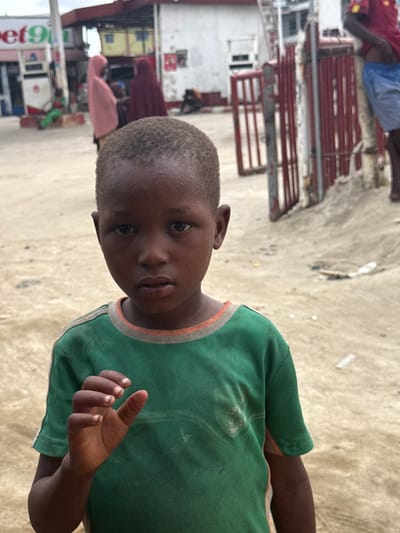 A young Black boy with short, dark hair and dark eyes stands in the foreground, looking directly at the camera with a somber expression. He is wearing a green t-shirt with orange and gray trim around the neckline and sleeve cuffs. His right hand is raised, with his thumb and index finger pinched together in a small gesture.
The background suggests an urban environment, likely a street or open area. To the left, a gas station with a white and red sign that partially reads "bet9" is visible, with gas pumps and a canopy. Several people are in the background, including two women wearing headscarves (one pink, one maroon) near the gas station. Further back, there's a building with a white facade and a red, slatted gate. The ground is sandy or dusty, with some debris scattered around. The lighting suggests an overcast or cloudy day. The overall scene appears to be set in Lagos, Nigeria, given the visual context and common urban features.