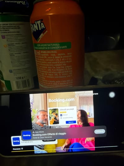 The image shows a smartphone screen displaying a Booking.com advertisement. The ad features a man and a woman with a beach in the background. The phone is placed in front of a shelf with various items, including a can of Fanta and a package of Rothmans cigarettes. The Fanta can has Italian text on it, and the advertisement on the phone is in Romanian and Italian. The text on the Fanta can indicates it is a product of Italy. The advertisement on the phone shows "Booking.com Offerte di viaggio" which translates to "Booking.com Travel Offers" and "Hotel & Case Vacanze" which translates to "Hotel & Holiday Homes." It also shows "Căsuță pe plajă" which translates to "Beach House." Below this is a rating of "9.0 Minunat" (Wonderful) and "Acceptă animale" (Accepts animals). The ad also has a "Rezervă" (Reserve) button. The phone displays a banner at the bottom that reads "Реклама" which is Russian for "Advertisement". The presence of Italian text on the Fanta can and the Romanian and Italian text on the advertisement, along with the mention of Carpignano Salentino, Italy, suggests the scene is likely set in Italy, possibly in or near Carpignano Salentino, though the image itself does not provide direct visual evidence of this specific location. The scene is indoors, with artificial lighting.