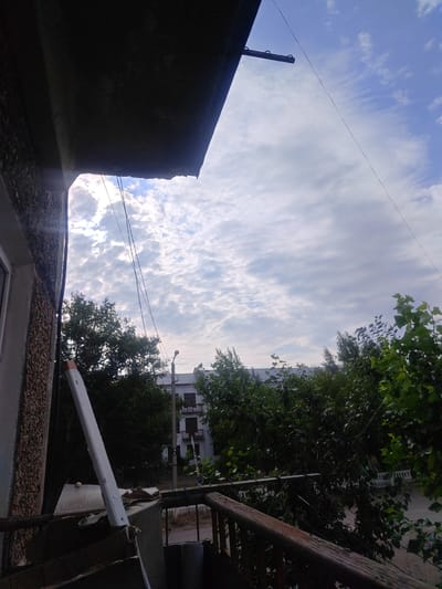 This media file depicts a view from a balcony, looking out towards a cloudy sky with a hint of blue peeking through. The sky is filled with scattered, wispy clouds, suggesting it might be mid-morning or late afternoon.
In the foreground, the edge of a balcony railing made of dark wood is visible. To the left, a rough-textured wall of a building is partially in frame, with a white object leaning against it, possibly a piece of construction material. Several thin wires are strung across the sky.
Beyond the balcony, lush green trees form a dense canopy. Behind the trees, a multi-story building with balconies and windows can be seen. A street lamp is also visible, and what appears to be a road or path runs between the trees and the building.
The overall impression is that of a calm, ordinary day in an urban or semi-urban setting. There are no people visible, and the scene does not suggest any specific activity or event. The lighting indicates it is daytime, and the cloudy sky hints at potentially variable weather. The location context provided is Ereymentaw, Kazakhstan. There is no visible text in the image.