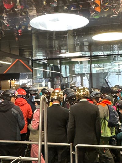 The image, if rotated 90 degrees counter-clockwise to be upright, presents an indoor scene within what appears to be a busy winter sports equipment store or rental facility. The overall lighting is bright, with strong reflections.
Several individuals are visible, primarily from behind, suggesting they are browsing or interacting with the displayed gear. In the mid-ground, a prominent figure wears a black winter jacket and a distinctive, shiny gold-colored helmet that appears to be textured or studded. This person's reflection, wearing an identical jacket and helmet, is clearly visible in a large, highly reflective surface (likely a mirror or polished wall) that occupies the left side of the frame. This reflective surface also captures the reflections of multiple large, circular, bright overhead lights.
To the right and slightly in front of these gold-helmeted figures, other customers are present. One person wears an orange or red winter jacket and a black helmet. Another, further down and to the right, wears a dark jacket and a black helmet, with the partial brand name "OAKLEY" faintly visible on the helmet's side. Near them, a person in a pink jacket is also visible. In the upper right corner, a grey helmet with the possible brand name "LOVE" or "LOVA" is seen on a rack, along with other colorful helmets and jackets (green, blue).
A wide variety of helmets, mostly black but also in colors like orange, green, and gold, are arranged on shelves and racks throughout the facility. In the foreground on the far right, a silver metal railing is present, possibly part of a queue barrier or display fixture. Through large windows or glass partitions in the background, a bright, somewhat indistinct outdoor or distant indoor area with structural elements can be seen, hinting at a larger complex or an external view.
The activity depicted is consistent with people selecting or trying on winter sports helmets and related attire, possibly preparing for skiing, snowboarding, or other cold-weather activities. The presence of numerous helmets and winter jackets indicates a focus on specialized gear.