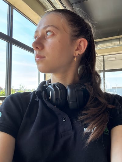 A young woman with dark hair, wearing a black polo shirt and black headphones, is shown in a close-up, side profile. She has light skin and light-colored eyes, and is wearing a small gold earring in her left ear. Her hair is pulled back, with some strands loose around her face. The headphones are on her head, positioned over her right ear.
The background suggests an indoor setting with large windows that offer a view of a bright, sunny sky and some greenery outside, indicating it is daytime. The scene appears to be a modern, well-lit space, possibly an office or a cafe, with industrial-style ceiling fixtures visible. The overall mood is relaxed and casual. There is visible text on the woman's shirt, which reads "Nigra" and "Bavaria" in green lettering. The location context is Elsenfeld, Germany.