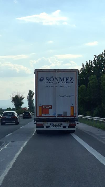 The image shows the rear view of a white semi-trailer truck with the logo and text "SÖNMEZ BUSTAS & LOJISTIK" on its side. The truck has a Turkish license plate "16 AOY 878". The truck is on a highway, with other cars visible in the background. The sky is cloudy, and there are trees and greenery on the side of the road. The scene appears to be a daylight shot taken from another vehicle on the highway. The location is not Benkovski, Bulgaria, as the license plate is Turkish and the text on the truck is in Turkish.