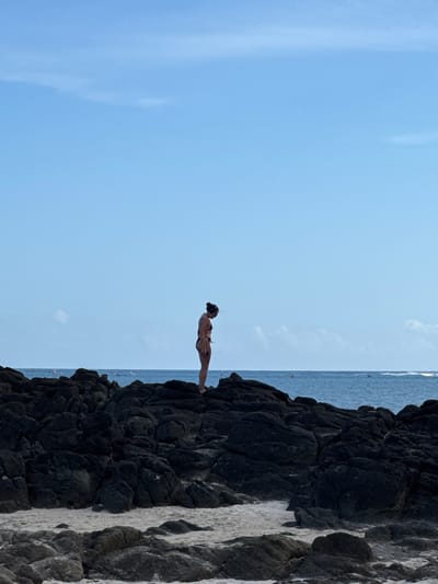 A vertically oriented photograph shows a woman in a bikini poised on the edge of a dark, rocky cliff ready to jump into the sea in Selongbelanak, Indonesia. The sky is pale blue with scattered clouds. The ocean is visible as a thin strip along the top, and several small boats are also visible in the sea, indicating human activity.