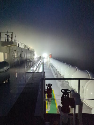 The image depicts a vessel at night, likely at sea or in port, given the presence of lifeboats lined up along the side and industrial piping. The scene is dimly lit, suggesting nighttime, and a thick fog or mist obscures the background, creating a hazy, ethereal atmosphere. The fog diffuses the light, causing a bright glow in the distance, possibly from shore or other vessels.

Prominently featured are a series of white, enclosed lifeboats, their rounded forms reflecting the limited light. Below the lifeboats, industrial elements are visible, including metal railings, pipes, and valves. Two red valves with black wheels are particularly noticeable in the foreground. One lifeboat, colored orange and red, is also visible, seemingly deployed or ready for deployment.

The perspective is from a low angle, looking upwards and along the length of the vessel. The surface of the ship appears wet, reflecting the ambient light and contributing to the overall moody ambiance. The metallic surfaces and industrial equipment suggest a working vessel, possibly a cargo ship or a research vessel.

The overall mood is one of quiet isolation and industrial utility, amplified by the obscuring fog and the late hour. There is no visible text in the image. The location context of Hitachinaka, Japan, doesn't offer specific visual cues within the image itself, but it places the scene within a coastal industrial area.