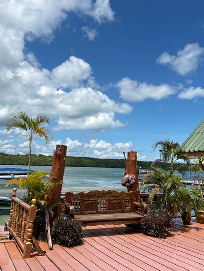 This is a landscape-oriented, slightly tilted, medium-shot photograph of a wooden deck overlooking a body of water. The sky is a vibrant blue with scattered white, fluffy clouds. A section of a green corrugated metal roof is visible in the upper left corner.

The deck itself is made of reddish-brown wooden planks. Several potted plants, including palm trees and other foliage, are arranged on the deck, suggesting an outdoor setting. A unique, rustic bar or counter made of rough-hewn wood and intricately carved panels is a prominent object. Another wooden post stands vertically to the left of the bar.

In the background, beyond the deck, is a large expanse of calm, light blue water. A lush, green, tree-covered coastline forms the horizon. Small boats with blue canopies are docked in the water below the deck.

The lighting suggests it is daytime, likely late morning or early afternoon, given the brightness and the shadows cast by the plants and structures. The weather appears to be clear and sunny, with a pleasant atmosphere implied by the tropical vegetation and the serene water. There is no discernible activity or event taking place, and no people are visible. The overall impression is one of a tranquil tropical location, possibly a restaurant or bar with a view. No text is visible in the image. The location context provided is Tambon Ko Panyi, Thailand.