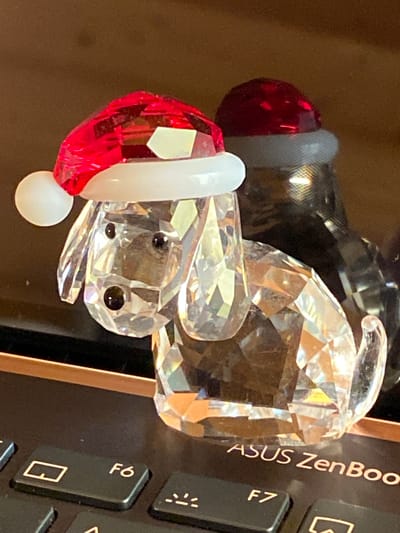 The image shows a close-up, slightly blurry shot of a crystal figurine of a mouse wearing a Santa hat. The figurine is made of clear crystal with faceted surfaces that catch the light, giving it a sparkling appearance. The mouse has two small black eyes and a red and white Santa hat. The figurine is positioned on what appears to be a laptop keyboard. The keys are black, and the text "ASUS ZenBook" is visible vertically along the side of the laptop. There is also a key labeled "F7" visible. The background is a blurred, warm brown color, possibly a table or desk. The lighting suggests it's indoors, possibly during the day as there are bright reflections on the crystal. The overall scene is a still life, highlighting the decorative crystal figurine.