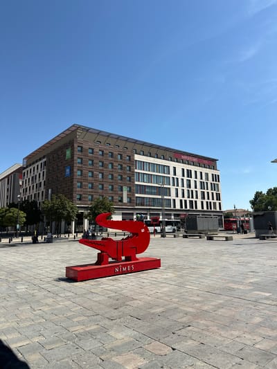 The image depicts a bright, sunny day in Nîmes, France, with a clear blue sky. In the foreground, a large, bright red sculpture of a crocodile is prominently displayed on a stone-paved plaza. The sculpture is a stylized representation of a crocodile, with sharp angles and a flat, geometric design. The word "NÎMES" is clearly visible in white lettering on the base of the sculpture.

Behind the sculpture, a modern, multi-story building dominates the background. This building appears to be a hotel, as indicated by the "ibis" and "ibis budget" logos visible on its facade. The building features a mix of brown and white cladding, with numerous windows arranged in a grid pattern. Some windows have colorful accents. The top of the building is capped with a slatted roof, possibly incorporating solar panels.

To the left of the building, another building with a lighter facade and vertical accents is partially visible. The plaza itself is paved with large, square stone tiles, and several trees are planted in rows along the edge of the plaza and surrounding the buildings. In the mid-ground, there are benches and what appear to be street furniture or kiosks. Several people are visible in the distance, walking and sitting on benches. Two red buses are parked in front of the building on the right side of the image, and a white bus is also visible on the left. The overall scene suggests a public space in a city, likely a transportation hub or a central square.