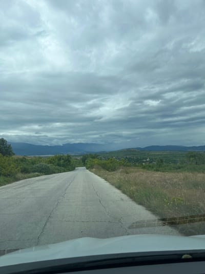 The image shows a cracked asphalt road leading into the distance towards a mountainous landscape under a cloudy sky. The road is flanked by dry grass and green foliage. In the distance, a village with buildings and a prominent church spire is nestled among rolling green hills, with a range of mountains forming the backdrop. The overall impression is that of a rural scene in Bulgaria, likely near the town of Pamidovo, given its location within a valley surrounded by mountains. There are no people or animals visible in the image. The perspective suggests the photo was taken from inside a vehicle, looking forward through the windshield.