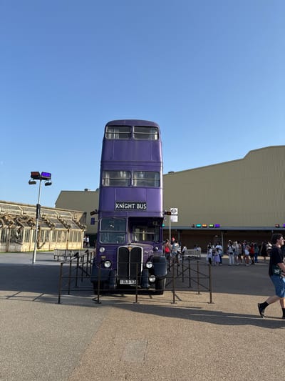 This image captures a scene featuring the iconic purple Knight Bus, a prop from the Harry Potter franchise, parked outdoors. The bus, a double-decker vehicle, is prominently positioned in the foreground, with its "KNIGHT BUS" sign clearly visible on its side. Behind the bus, a line of people, likely visitors or fans, are queuing, suggesting an attraction or event.
The setting appears to be an outdoor area with a paved ground and a large building in the background. The building has a ribbed, light-colored facade and a row of windows with colorful lights above them, hinting at a themed environment, possibly a theme park or a movie set recreation. There's also a glimpse of another building with ornate architectural details to the right.
The sky is a clear, bright blue, indicating a sunny day with good weather. The lighting suggests it is daytime, likely mid-morning or afternoon, given the strong shadows cast by the bus and the people. A traffic light-like structure with multiple colored lights is visible in the lower left quadrant, adding to the urban or attraction-like feel of the location.
The image is oriented sideways, with the bus and people appearing upright when the image is rotated. The license plate on the front of the bus reads "OLD 717". The overall mood is one of anticipation and enjoyment, as people are gathered to experience something related to the Harry Potter universe. The presence of the Knight Bus strongly suggests a location associated with the Harry Potter films, such as a theme park in Orlando or a studio tour in the UK. Given the "Three Rivers, United Kingdom" location context, this is likely Warner Bros. Studio Tour London – The Making of Harry Potter, which features the Knight Bus.
