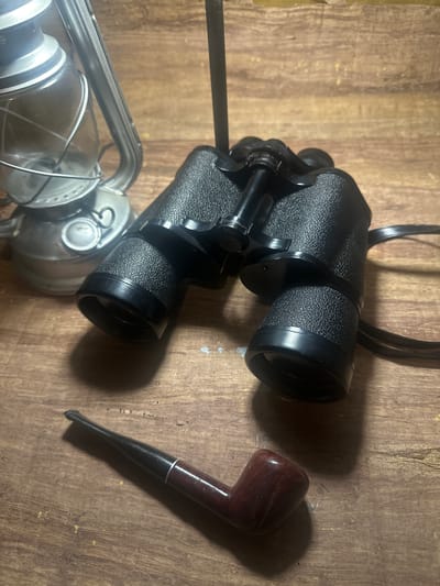 A still life composition is set on a weathered wooden surface. On the left side of the frame sits a vintage-style oil lantern with a silver metal frame and a clear glass chimney. To the right of the lantern are a pair of black binoculars with a textured grip. In the foreground, positioned diagonally from lower left to upper right, is a dark wood smoking pipe with a dark stem and a silver band around the neck. The background is a blurred wooden texture, suggesting an indoor or rustic outdoor setting. The lighting is dim, casting shadows from the objects onto the wooden surface. The image does not depict any specific location in the city of Lapės, Lithuania, or any people or actions.