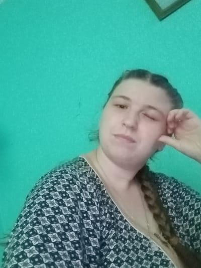 A woman is taking a selfie in front of a teal wall in Kitskany, Moldova. She has her right hand near her face, and her left eye is closed. Her hair is in a braid that falls over her left shoulder. She is wearing a patterned blouse. There is a picture frame visible in the top right corner of the image.