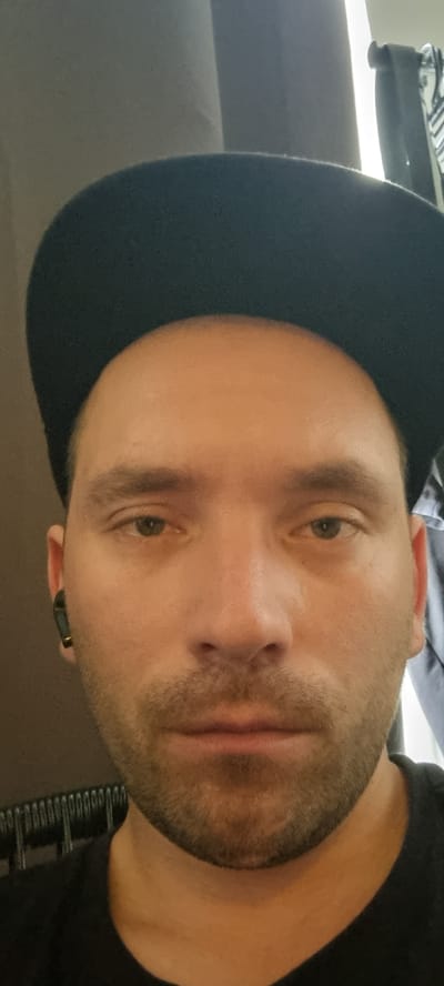 The media file is a close-up, horizontally oriented, over-the-shoulder shot of a person's face, likely a man, captured indoors. The lighting suggests it is daytime, possibly late afternoon or early evening, given the warm, slightly diffused light. The scene is indistinct, but appears to be a domestic or personal setting, with hints of what could be a shelf or rack in the background.
The individual is wearing a dark baseball cap, tilted forward. A black t-shirt is visible around the neck and shoulder area. The person has short, light brown hair, stubble on their face, and is wearing a black wireless earbud in their right ear. The dominant feature is their face, with visible skin texture, a closed mouth, and light brown eyes looking slightly off-camera. There is a slight reddish hue on the bridge of their nose.
The image is somewhat blurry and the orientation is off, suggesting it was taken by the subject themselves. The overall mood is casual and candid, with no specific activity or event clearly depicted. There is no visible text in the image. The location context of Haarlem, Netherlands is not directly evident from the visual cues within the image itself.