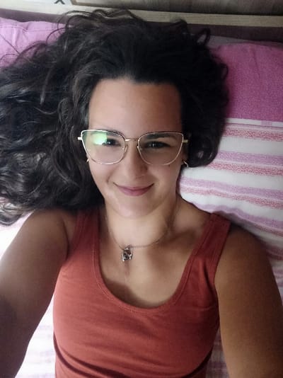 A female individual is depicted in a close-up, high-angle shot, lying on her back in an indoor setting, likely a bedroom, in San Marzano sul Sarno, Italy. She has long, dark, wavy hair spread out on a solid pink pillow. She wears gold-framed, cat-eye style eyeglasses and a rust-colored sleeveless top. A thin, silver-colored necklace with a dark pendant is visible around her neck. Her face is clear, with a slight smile, and her brown eyes are directed towards the camera. The background consists of the pink pillow and white bedsheets with faint pink stripes.