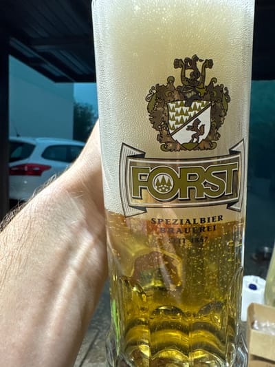 The image shows a close-up of a glass beer bottle, likely taken outdoors during the day. The bottle is filled with golden beer and has a thick layer of foam at the top. The label on the bottle features a crest with an animal and shield design, and the word "FORST" prominently displayed in large, stylized letters. Below "FORST," smaller text reads "SPEZIALBIER BRAUEREI SEIT 1857." A person's arm is visible in the foreground, partially obscuring the lower part of the bottle and the background, which appears to be a street scene with a white vehicle and some buildings. The lighting suggests it is daytime, but the specific time is not evident. There are no strong emotional cues conveyed, and the focus is primarily on the beer and its branding. The location context of Sant'Angelo di Piove di Sacco, Italy, is provided, but not visually discernible from the image itself.