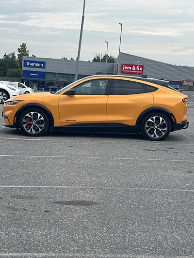 In Slitu, Norway, an orange Ford Mustang Mach-E is parked in a lot in front of two stores. The store to the left has a blue sign that says "thansen" and "Velkommen" below it. The store to the right has a red sign that says "jem & fix LAVPRIS BYGGEMARKED". The sky is overcast.