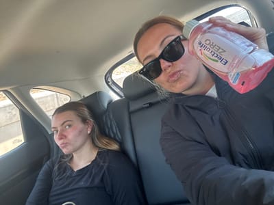 Two young women sit in the back of a car. One woman, wearing black sunglasses and a black jacket, holds up a bottle of Electrolyte Zero drink. Both women are pursing their lips. The woman seated near the car door wears a black shirt. A small earring is visible. The car's interior shows black leather seats and a light-colored ceiling. Outside the car window, trees and a concrete wall can be seen.