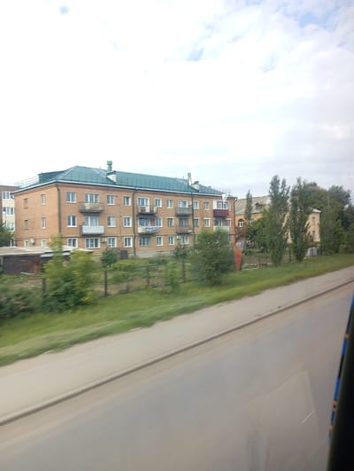 The image shows a residential building in Sol-Iletsk, Russia, on a cloudy day. The building is a multi-story brick structure with a green roof. It has numerous windows and balconies. In front of the building, there is a fence and some trees and grass. A road is visible in the foreground, with a car or bus passing by, as indicated by the blurry foreground. The scene appears to be a typical urban or suburban setting.