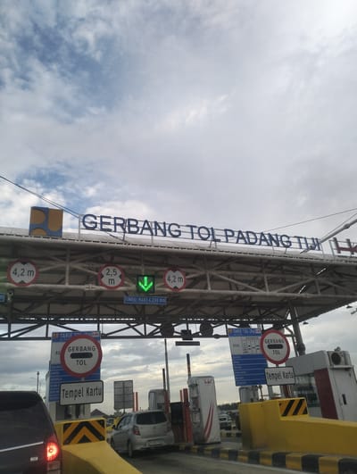 This media file shows a view of a toll gate on a cloudy day. The main subjects are the toll gate structure, vehicles, and signage. The toll gate is a large overhead structure with metal supports and a roof, displaying prominent signage.

The text on the overhead sign reads "GERBANG TOL PADANG TIJI H". Below this, there are circular signs indicating height limits: "4,2 m" and "2,5 m". A green downward-pointing arrow is also visible, indicating an open lane. Another sign below reads "TINGGI MAKS 4.200 mm".

On the right side of the road, there are toll booths and barriers. Signs on the booths include "TARIF TOL (RP)" and "GERBANG TOL". One sign instructs "Tempel Kartu", meaning "Tap Card". A list of locations is visible on a blue sign, likely indicating origins or destinations for toll pricing.

In the foreground, parts of cars are visible, one black car on the left and a silver hatchback on the right, suggesting traffic is moving through the toll. The overall scene is a typical toll plaza environment. The weather appears overcast with a mix of clouds and some blue sky visible. The location is identified as Pante Cermen, Indonesia. No people are clearly visible, but their presence is implied by the presence of vehicles and the function of the toll gate. The mood is functional and neutral, depicting a common travel infrastructure.