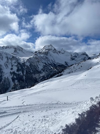 The image displays a vast, snow-covered mountainous landscape situated in Puy-Saint-André, France, under a partially cloudy blue sky. The foreground is dominated by an extensive field of fresh white snow, marked with numerous visible ski or snowboard tracks creating curvilinear patterns across the surface. A single grey metallic fence post, with a thin wire or cable attached, is partially buried in the snow on the lower left side. The midground reveals a series of snow-covered slopes descending into a valley, sparsely dotted with dark green coniferous trees. Further in the background, rugged, snow-capped mountain peaks rise prominently against the horizon. The sky exhibits patches of clear blue interspersed with scattered, fluffy white cumulus clouds. A small, dark, unidentified airborne object, possibly an aircraft or bird, is visible against the sky above the mountain range in the upper right portion of the frame. No human subjects are present, and no active interactions are depicted within the scene. The environment indicates a cold, high-altitude setting with ample snow cover.