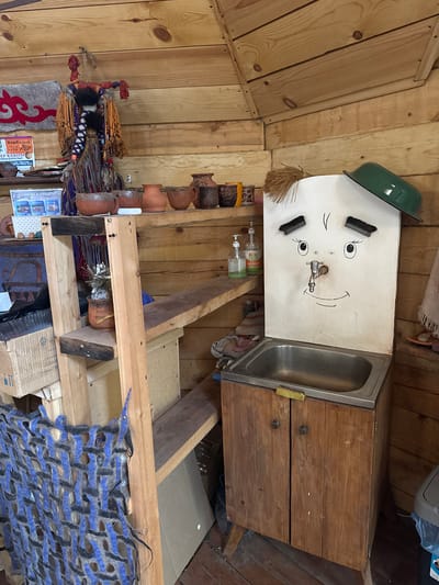 The image shows an interior scene, likely a rustic cabin or workshop, in what appears to be Askat, Russia, given the presence of Russian language signage. The walls are made of wooden planks. The focal point is a makeshift washbasin featuring a metal sink atop a wooden cabinet with two drawers. The basin has a white painted front, humorously decorated with drawn-on facial features including eyes, eyebrows made of brushes, and a curved smile. A green metal bowl acts as a "hat" for the face. Below the washbasin, there is a wooden shelf system holding various items. These items include a collection of small terracotta pots, bottles of liquid soap, and various decorative objects, including a traditional-style decorative item hanging on the wall. A shelf below contains folded textiles and cardboard boxes. The overall impression is one of a simple, utilitarian space.