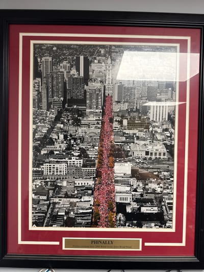 The image is a framed photograph of a parade in Philadelphia, Pennsylvania. The photograph is taken from an aerial perspective, looking down on a city street lined with buildings. A large crowd of people, dressed in red, are packed along the street, suggesting a celebratory event. The sky is overcast. Below the photograph is a brass plaque with the inscription "PHINALLY Phillies celebrate their 2008 World Series down Broad Street". The frame is black with a red mat.