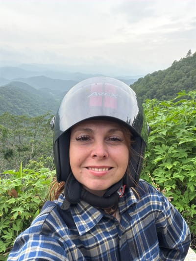 A woman wearing a black helmet and a blue plaid shirt smiles at the camera in front of a misty mountain landscape in Samoeng Tai, Thailand. The woman has red hair and is wearing makeup, including false eyelashes. The helmet has a red logo on it. The background consists of rolling green mountains covered in trees, with a hazy sky. Lush green foliage is visible in the foreground, framing the woman on the right side of the image.