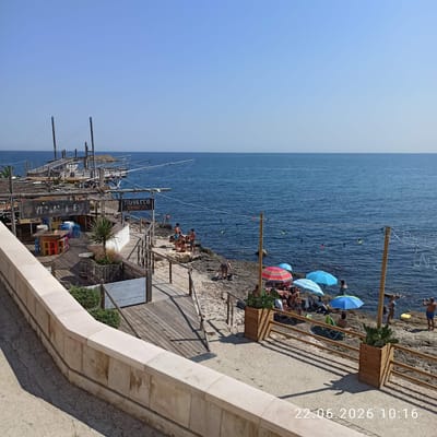 The image depicts a seaside scene in Trani, Italy, likely a tourist spot. A wooden structure, possibly a restaurant or beach club, is visible with tables, chairs, and umbrellas. Several people are present, some sunbathing on the rocky shore, others standing or walking. The sea is calm and blue, stretching to the horizon under a clear sky. The architecture of the wooden structure suggests a rustic, beachside ambiance. The date stamp at the bottom right indicates the photo was taken on June 22, 2026, at 10:16 AM.