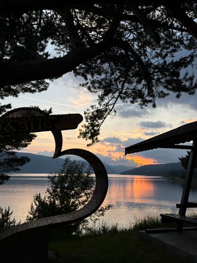 The image depicts a sunset scene over Dospat Dam in Dospat, Bulgaria. In the foreground, silhouetted elements frame the view. To the left, a large, dark tree branch extends from the top, beneath which a wooden sign is partially visible. The sign reads "КОМПЛЕКС ОАЗИС" (Complex Oasis) and "Стаи за гости" (Rooms for guests) in Cyrillic, and features a distinctive curved, abstract design. Below the sign, a dark, curved wooden structure, possibly a decorative bench, is present. Scattered smaller trees and bushes appear between these structures and the lake. To the right, a dark silhouette of a wooden structure with a slanted roof, resembling a gazebo or shelter, and part of a wooden bench are visible. Pine branches are also visible at the top right. The midground is dominated by the calm waters of the lake, which intensely reflect the vibrant orange, red, and yellow hues of the setting sun. Distant mountain ranges form a dark silhouette against the horizon. The sky above features a gradient of sunset colors near the horizon, transitioning to muted blues and greys higher up, with scattered clouds and several discernible airplane contrails. No individuals are present in the scene.