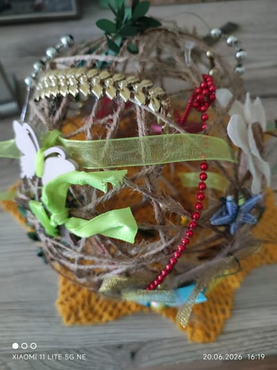 The image displays a close-up, slightly angled view of a decorative spherical object. This object is constructed from twisted twine and adorned with various embellishments, including gold-colored leaf-shaped sequins, strings of red beads, and white and blue decorative butterflies. Green ribbons are also wrapped around the sphere. The object rests on a yellow crocheted mat. The background consists of a wooden surface and a blurred wall. The timestamp at the bottom of the image indicates "20.06.2026 16:19" and "XIAOMI 11 LITE 5G NE". There is no visible indication of the city of Böblingen, Germany, or any specific environmental or contextual clues that would allow for such a determination.