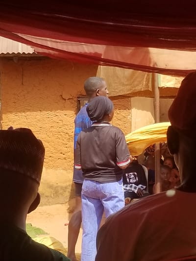 This image depicts an outdoor gathering in Ochadam, Nigeria. In the immediate foreground, the backs of two individuals' heads and shoulders are visible; the left individual wears a dark cap. In the midground, a dark-skinned female stands facing slightly right, wearing a black head covering, a dark short-sleeved t-shirt with red and white striped cuffs, and light blue denim jeans. Behind her to the left, a dark-skinned male in a blue t-shirt is partially visible. The background features a building with textured, light brown earth walls and a corrugated metal roof. Red and beige translucent fabrics are draped overhead, forming a temporary canopy or shading structure. To the right of the midground subjects, a group of individuals, including children, are partially visible; one child gazes towards the camera. The ground is bare, light-colored earth, and the scene is brightly illuminated by direct sunlight.