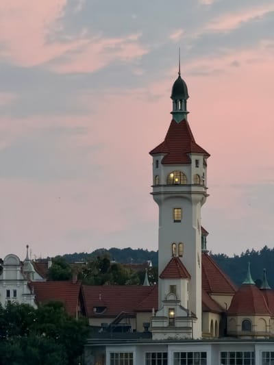 The image depicts a view of Sopot's historic Lighthouse and the surrounding architecture at dusk. The central focus is the white lighthouse with its red-tiled conical roof and a prominent spire, featuring several windows, some of which are illuminated, indicating an interior light source. The lighthouse is a prominent structure within a complex of similarly styled buildings, characterized by red tile roofs and white facades. Lush green trees are visible in the foreground and interspersed among the buildings, suggesting a verdant environment. In the background, a dense line of trees forms a dark silhouette against the horizon, with a gradient sky transitioning from pale pink to a soft blue, suggesting the sunset. The overall impression is one of a serene, historical urban landscape at the end of the day.