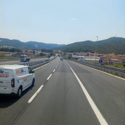 This is a medium-angle, eye-level shot captured on a sunny day on a highway in Quiliano, Italy. The highway has multiple lanes and is bordered by metal guardrails. In the foreground, a white Ford Transit van with "AmicoBlu" branding is visible in the left lane. The van has a website address and phone number printed on its side. Further ahead on the highway, a white truck with a crane is in the right lane, and other vehicles are present. The background shows a hillside town with red-tiled roofs and green trees, and mountains in the distance. A blue sign with a white arrow and a triangular yield sign are also visible.