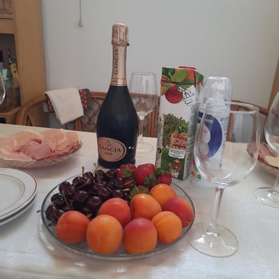 A table is set for a meal with a bottle of Gancia Asti champagne, two wine glasses, a carton of juice, a dish of sliced ham and cheese, a plate of cherries and apricots, and a plate of strawberries. The scene appears to be indoors, possibly on a patio or in a dining room, with a white tablecloth covering the table. The background includes a wicker chair and a white wall. The lighting is natural, suggesting daytime. No people are visible in the image. The location is not discernible from the image itself.